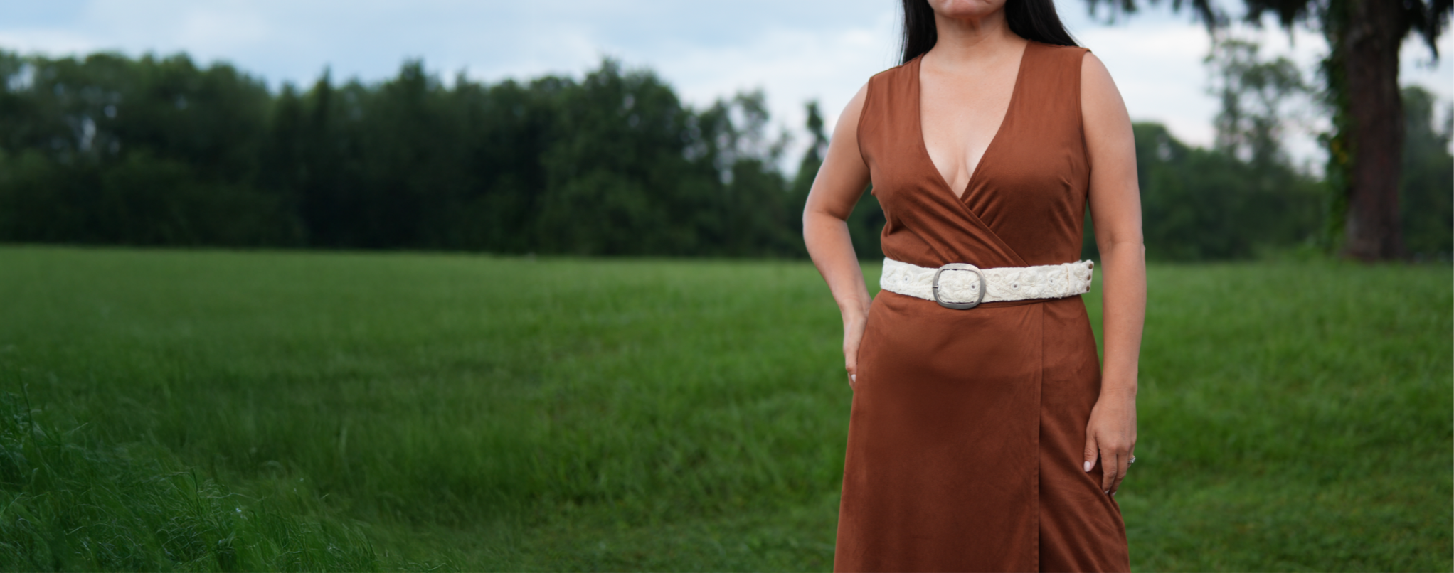 Woman in a brown dress standing in a grassy field displaying an embroidered Peruvian artisanal belt