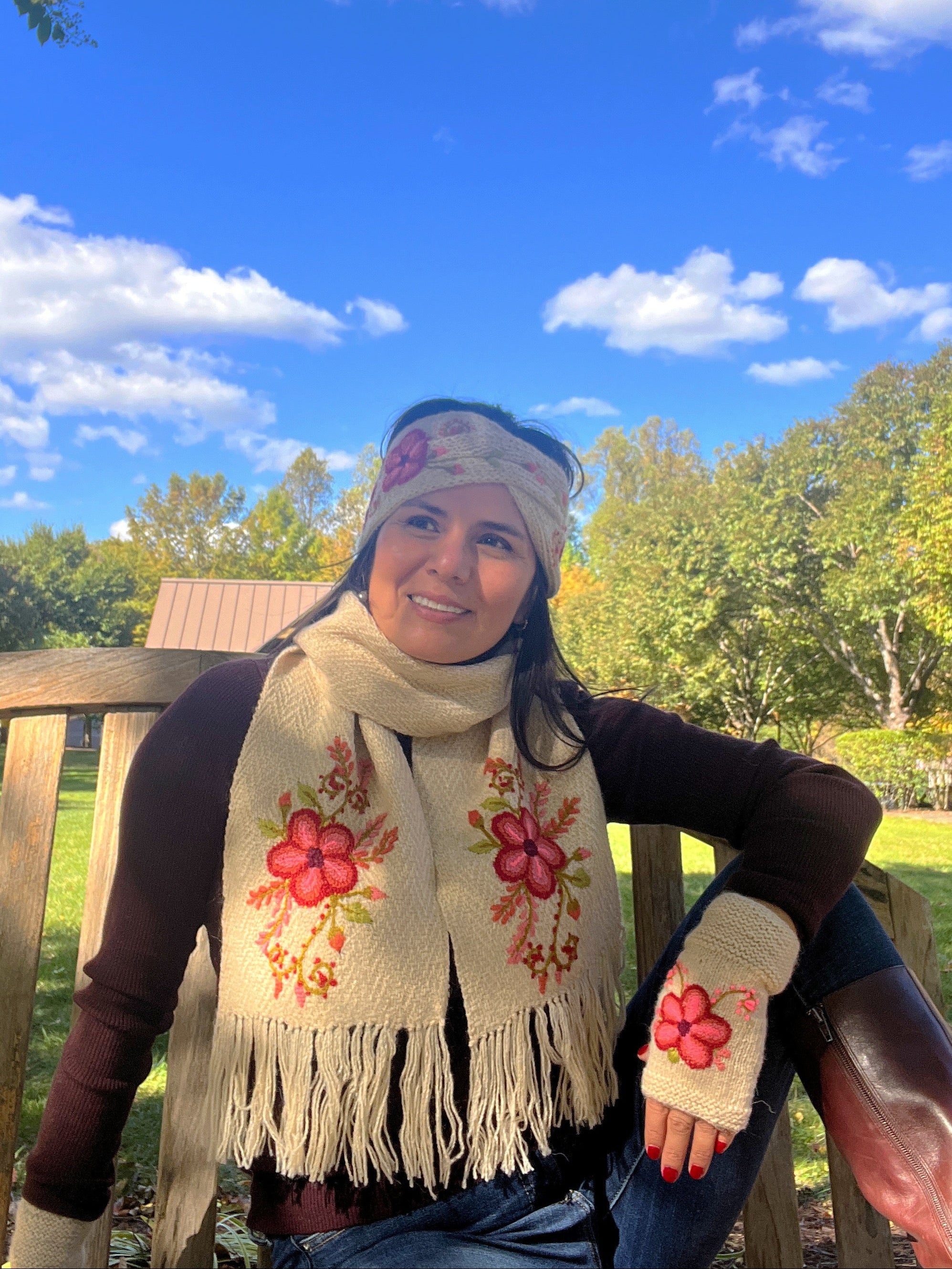 Woman sitting on a bench outdoors wearing a floral scarf and mittens, with a blue sky and trees in the background.