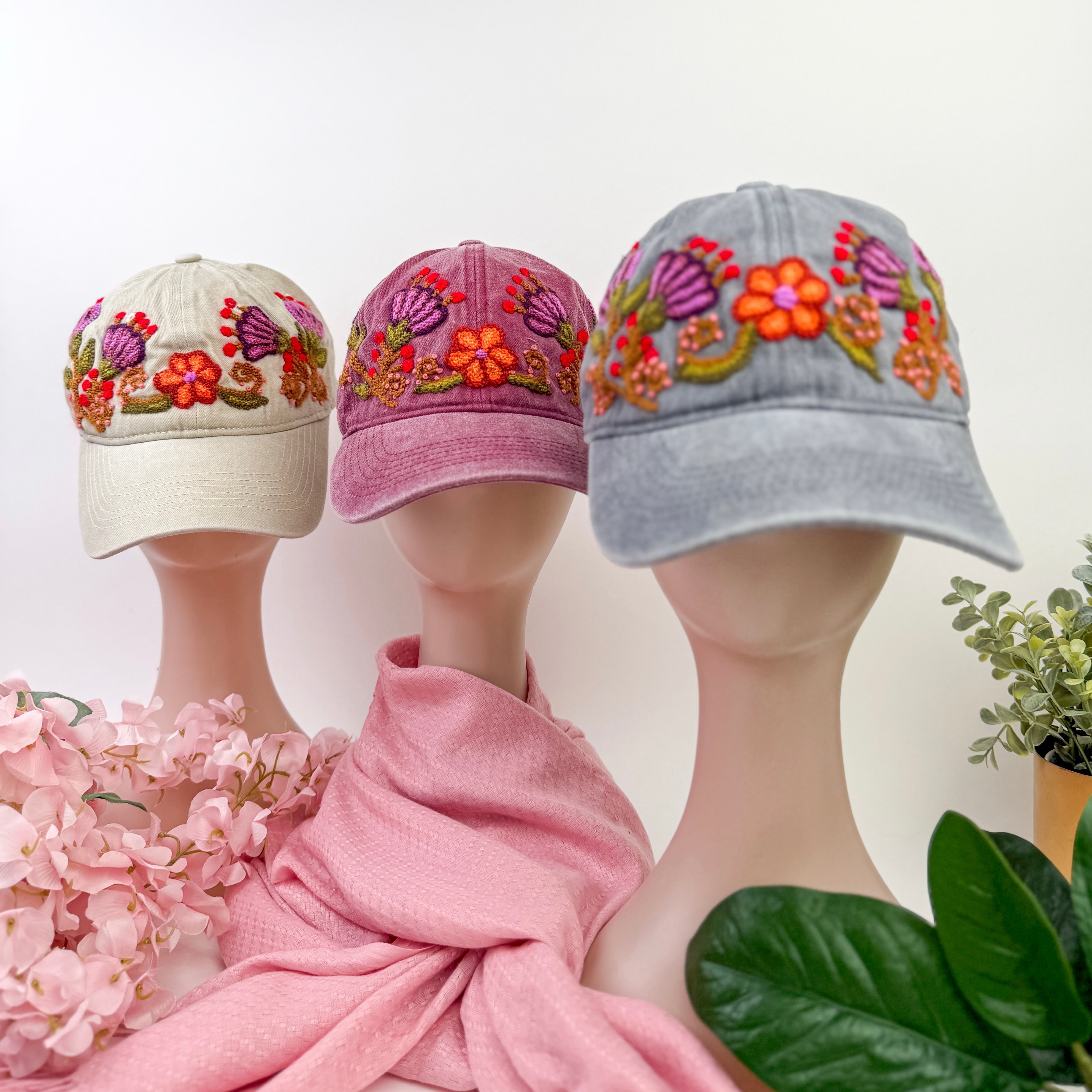 Three embroidered caps on mannequins with a pink scarf and plant in the background